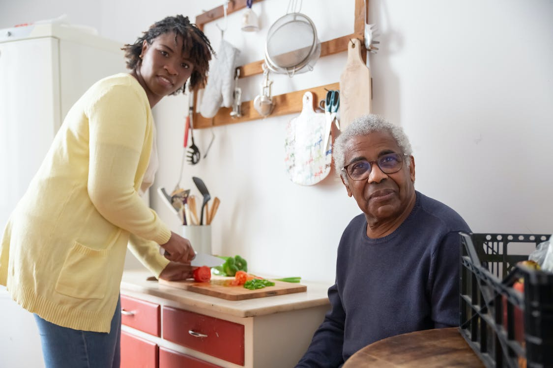 A caregiver is preparing food in the kitchen while an elderly man sits nearby.