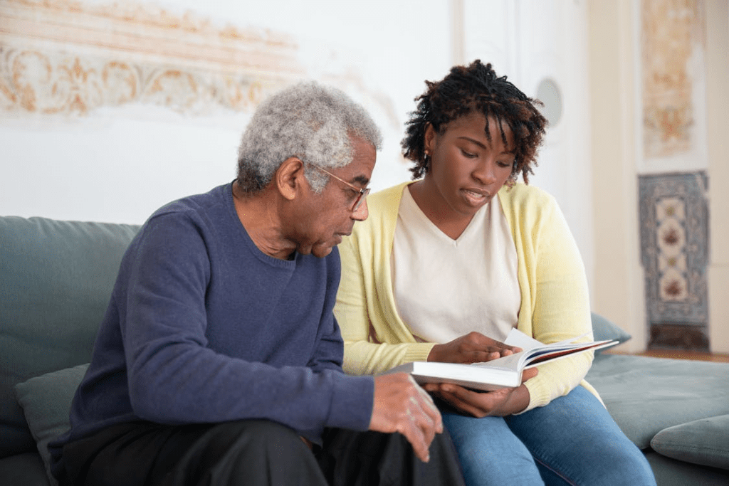 A caregiver is reading to an elderly man while both are sitting on a couch.