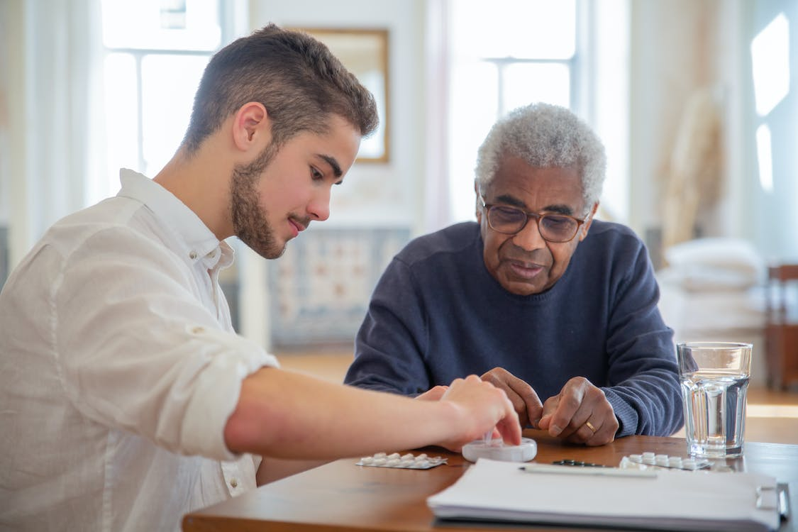 An elderly man is sitting at a desk with his caretaker as his blood pressure is being measured.