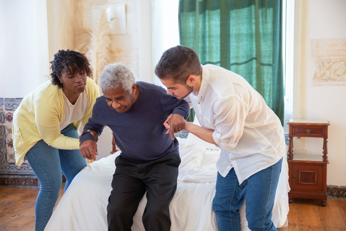 Two caregivers are helping an elderly man rise from bed.