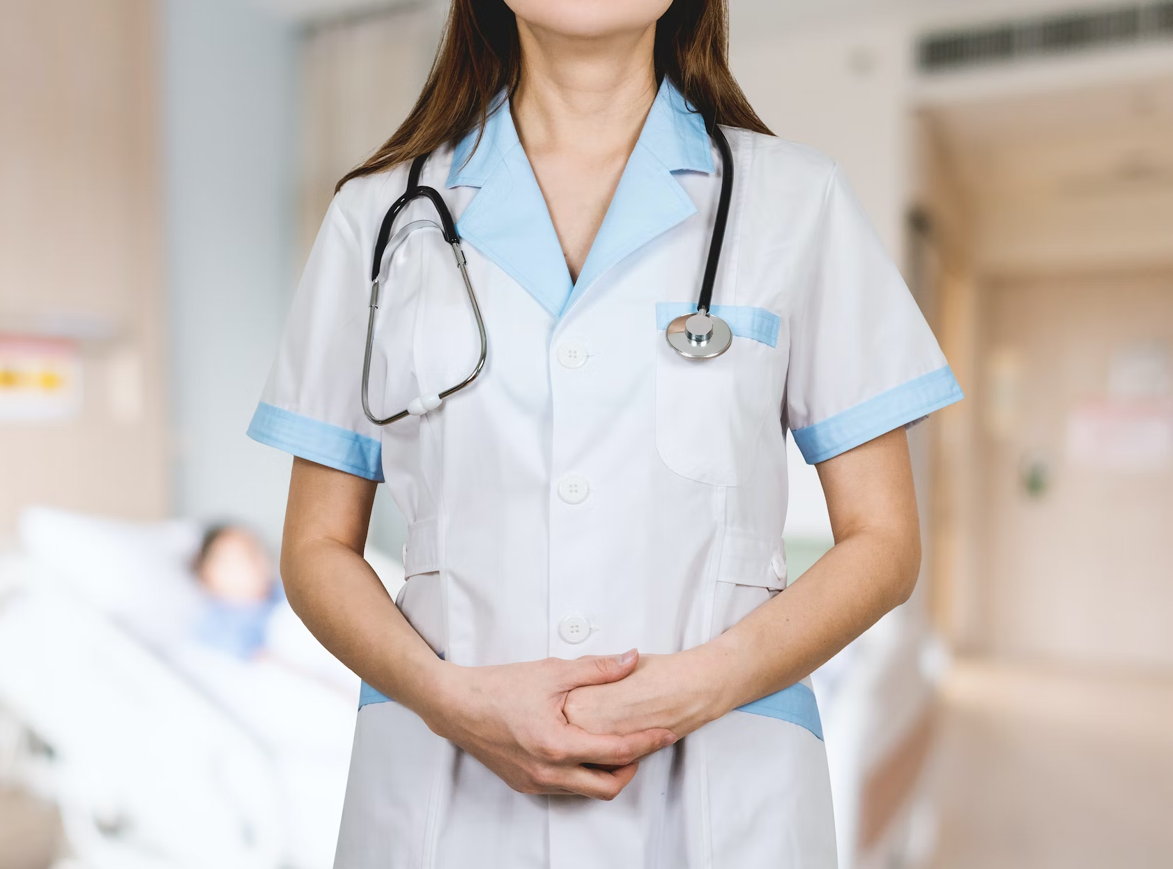 A woman in a nurse uniform and stethoscope stands near a bed.